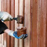 A person is applying brown paint to a wooden fence using a brush. They wear gloves to protect their hands and work diligently in a sunny outdoor setting.