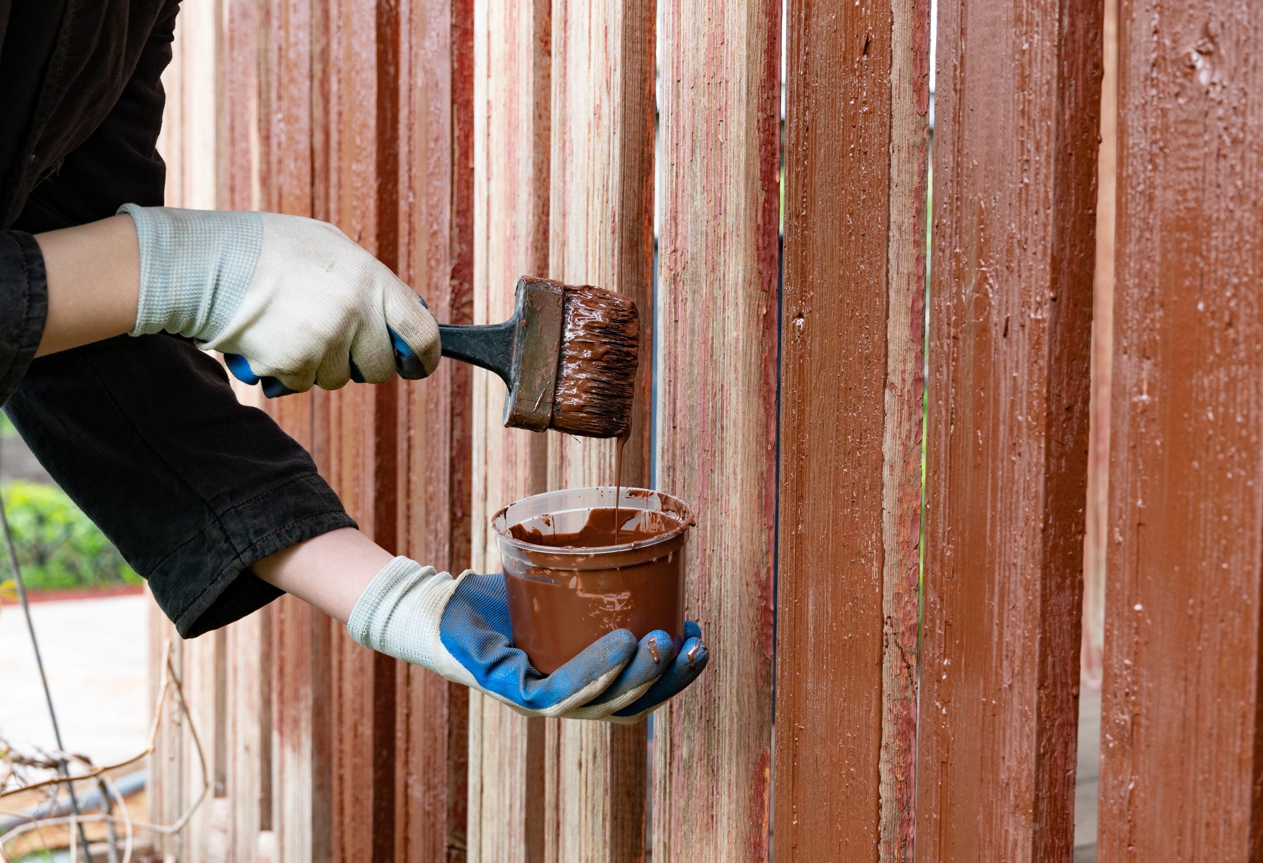 A person is applying brown paint to a wooden fence using a brush. They wear gloves to protect their hands and work diligently in a sunny outdoor setting.